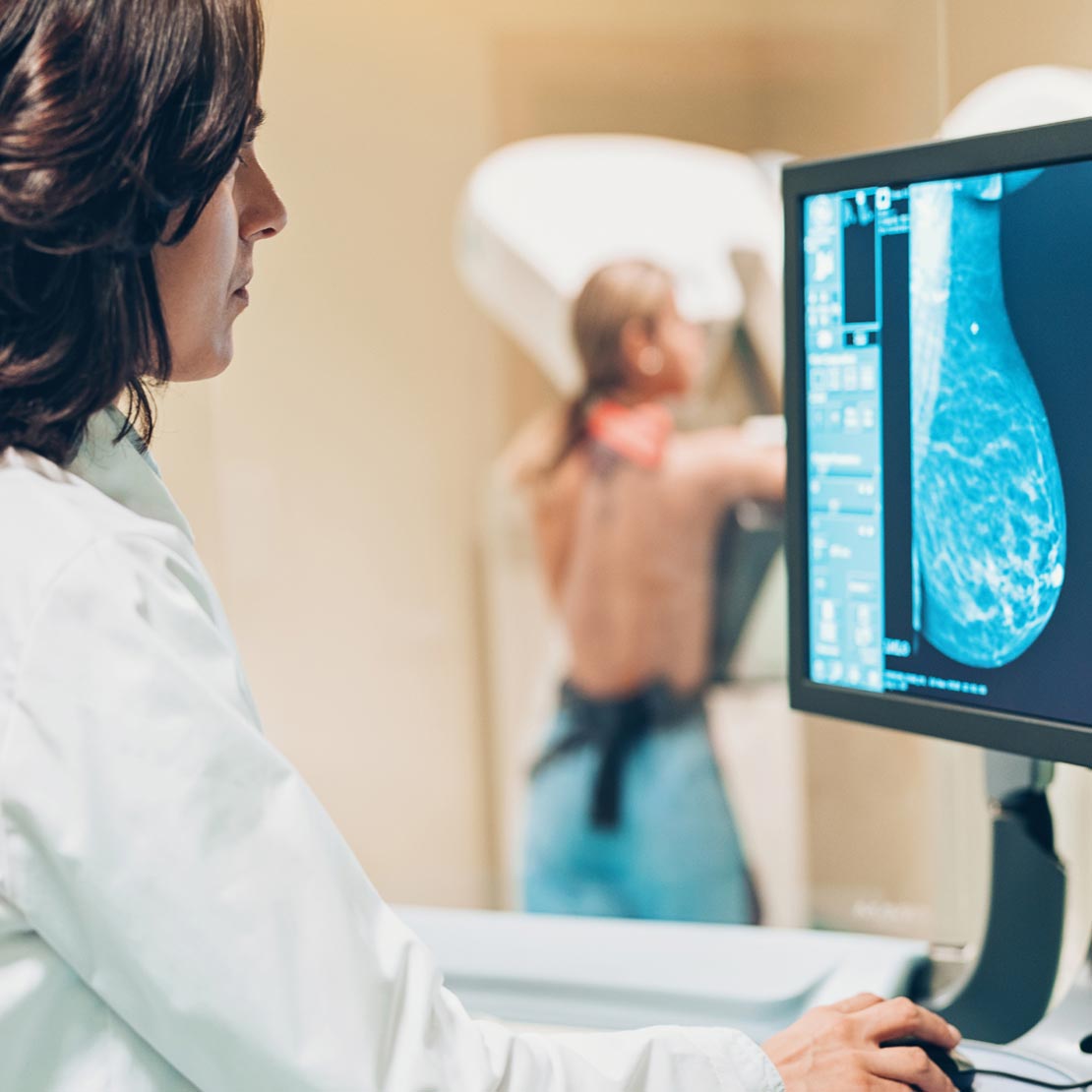A radiologist views a mammogram image on a large screen while a woman in the background is positioned at a mammogram machine.