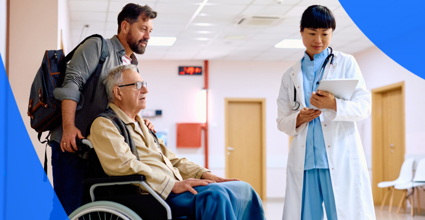 Middle-age man pushes elderly man in wheelchair down hospital hallway, stopping to review medical records in Ontario with health care professional.